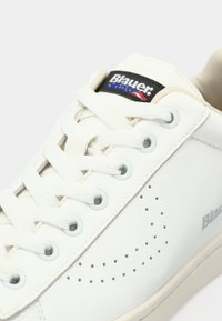 Λευκό δερμάτινο αθλητικό παπούτσι με διάτρητο σχέδιο, μπεζ λεπτομέρειες, στρογγυλά κορδόνια και λογότυπο στο γλώσσα με κόκκινα και μπλε στοιχεία.