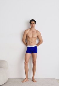 Boxer shorts en coton bleu avec une ceinture élastique blanche. Le modèle est debout pieds nus contre un arrière-plan neutre.