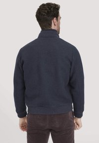 Man die met zijn rug naar de camera staat en een donkerblauwe trui met geribbelde kraag en manchetten draagt, gecombineerd met een bruine corduroy broek, tegen een effen achtergrond.