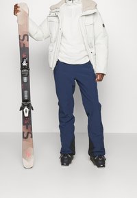 Skidutrustningen inkluderar en ljusfärgad vadderad jacka, ett vitt mellanlager, blå skidbyxor och svarta skidpjäxor som håller ett dekorerat par skidor med bindningar.