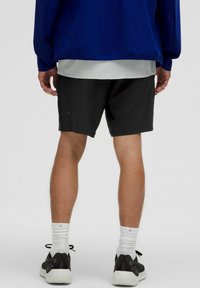Person stående vänd bort, klädd i svarta träningsshorts, vita strumpor, svarta sneakers, en vit tröja och en blå jacka.