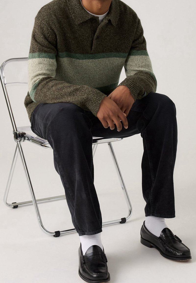 Homme assis sur une chaise pliante en métal, portant un pull rayé marron et vert, un pantalon noir, des chaussettes blanches et des mocassins noirs.