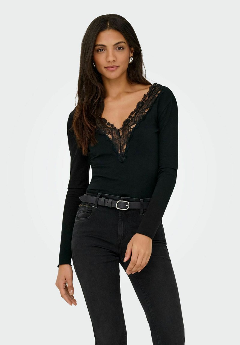 Zwarte met lange mouwen bodysuit met een diepe V-hals versierd met zwarte kant. Het model draagt het in een hoge taille zwarte jeans met een riem.