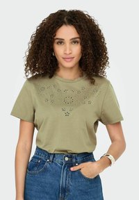 Olivenfarvet kortærmet t-shirt med blomsterudskæringer på forsiden, parret med højtaljede denimshorts.