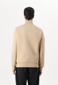 Beige sweatshirt med høy krage, lange ermer og ribbet midje, kombinert med mørke jeans. Enkel design, glatt tekstur.