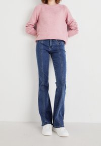 Light pink knitted sweater with a crew neck, paired with dark blue flared jeans featuring white stitching. White sneakers complete the outfit.