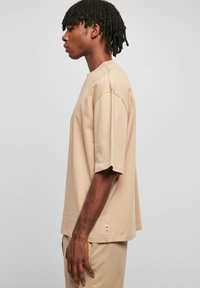 Tricou din bumbac bej, cu o croială relaxată, mâneci scurte și detalii subtile de cusătură. Prezintă un etichetă verde cu logo în partea de jos.