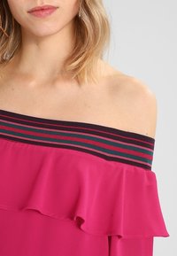 Top off-the-shoulder en fucsia con dobladillo con volantes, que presenta una banda negra con rayas de colores en verde, rosa y rojo oscuro.