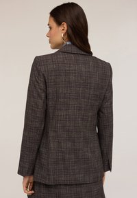 Blazer a quadri marroni con vestibilità su misura, dotato di un colletto classico, spalle strutturate e maniche lunghe, posto su uno sfondo chiaro.