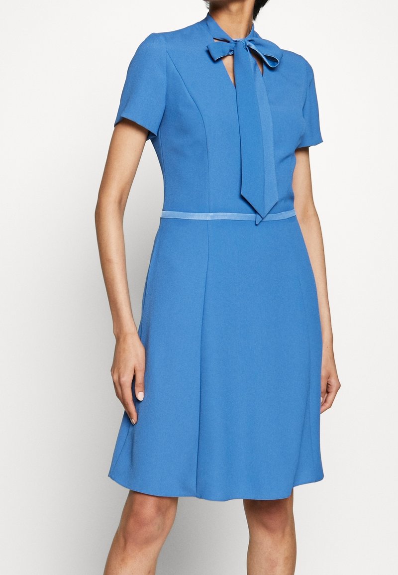 Femme portant une robe bleue longueur genoux avec des manches courtes et un nœud noué au cou, debout devant un fond clair uni.