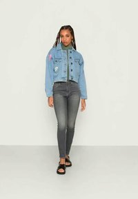 Jaqueta de ganga azul clara, cortada, com remendos, botões pretos, usada sobre um suéter de gola alta verde, acompanhada de jeans escuros e sandálias pretas.