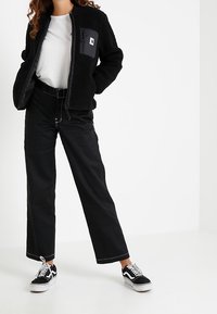 Zwarte fleece jas met een ritssluiting aan de voorkant en een borstzak, gecombineerd met wijde zwarte broeken en zwart-witte sneakers.