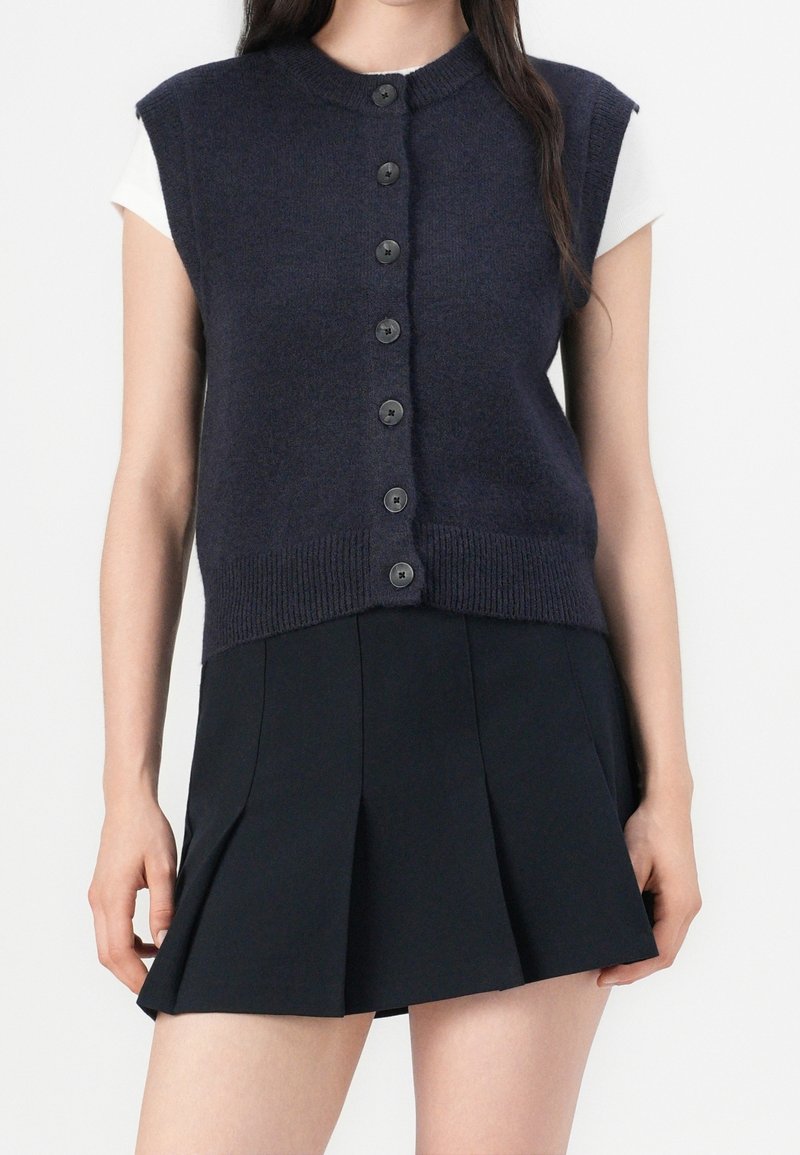 Femme portant un gilet sans manches bleu marine foncé boutonné sur un top blanc à manches courtes avec une mini-jupe plissée noire.