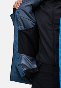Das Innenfutter der blauen und schwarzen Winterjacke besteht aus einem gestepp-ten grauen Innenfutter, mehreren Taschen und verstellbaren Ärmelbündchen mit Druckknöpfen. Weiche, glatte Texturen.