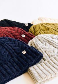 Set of five knit beanies in navy, burgundy, black, olive, and cream. Features textured cable patterns and ribbed cuffs. Labels on each beanie.