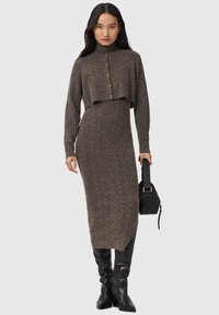 Pletené šaty tmavozelenej farby s rebrovanou textúrou, dlhými rukávmi a vysokým výstrihom, doplnené krátkym zapínacím svetríkom, v kombinácii s čiernou kabelkou a topánkami.