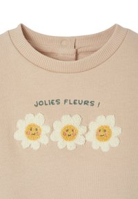 Helles rosa Sweatshirt mit drei gestickten weißen Gänseblümchen mit gelben Zentren und Gesichtszügen, beschriftet mit "JOLIES FLEURS !" in grün.