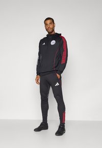 Schwarze Sport-Hoodie und Hose mit roten und weißen Streifen. Mit einem Logo auf der Brust, schlanker Schnitt und strukturiertem Stoff. Schwarze Schuhe.