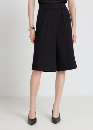 Donna che indossa culotte nere su misura fino al ginocchio abbinate a décolleté nere a punta e braccialetti dorati al polso sinistro.