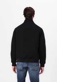 Schwarze Pulloverjacke mit hohem Kragen, Raglanärmeln und gerippten Bündchen, kombiniert mit blauen Jeans; schlichte Rückansicht vor einem weißen Hintergrund.