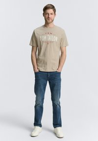 Beige katoenen t-shirt met rood en witte grafische tekst op de voorkant, gecombineerd met blauwe denim jeans en witte sneakers. Casual pasvorm en stijl.