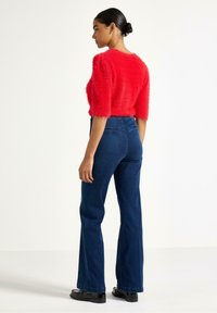 Maglione corto rosso peloso abbinato a jeans svasati blu scuro, indossato con scarpe nere. Presenta maniche corte e una vestibilità morbida.