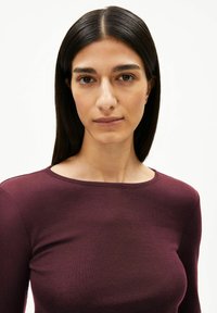 Bordeauxrotes Langarmshirt mit rundem Ausschnitt, gefertigt aus einem glatten, strukturierten Stoff, der eng am Körper anliegt. Minimalistisches Design, ohne sichtbare Muster.