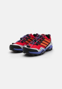 Rote und graue Trail-Schuhe mit schwarzen Akzenten, die orangefarbene Streifen, eine robuste Sohle, ein strukturiertes Mesh-Obermaterial und elastische Schnürsenkel mit Toggle bieten.