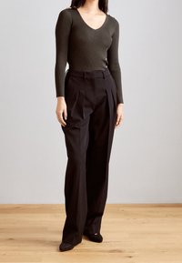 Mujer vestida con un suéter de canalé verde oliva oscuro y pantalones negros de pierna ancha de cintura alta, de pie sobre un suelo de madera.