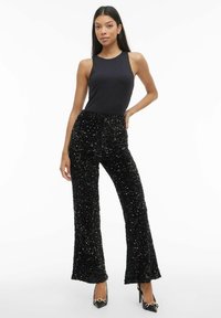 Ženska, oblečena v črno majico brez rokavov in široke črne hlače z visokim pasom, okrašene s šljokicami, ter črne visoke pete z zlatim verižnim detajlom.