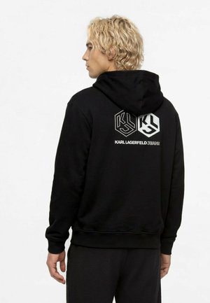 Muž s blond kučeravými vlasmi nosiaci čiernu mikinu s kapucňou s dvoma geometrickými logami a textom "KARL LAGERFELD JEANS" na chrbte.