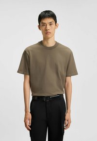 T-shirt in cotone marrone con scollo rotondo e maniche corte, caratterizzato da un sottile logo "HUGO" al centro. Indossato con pantaloni neri e una cintura.