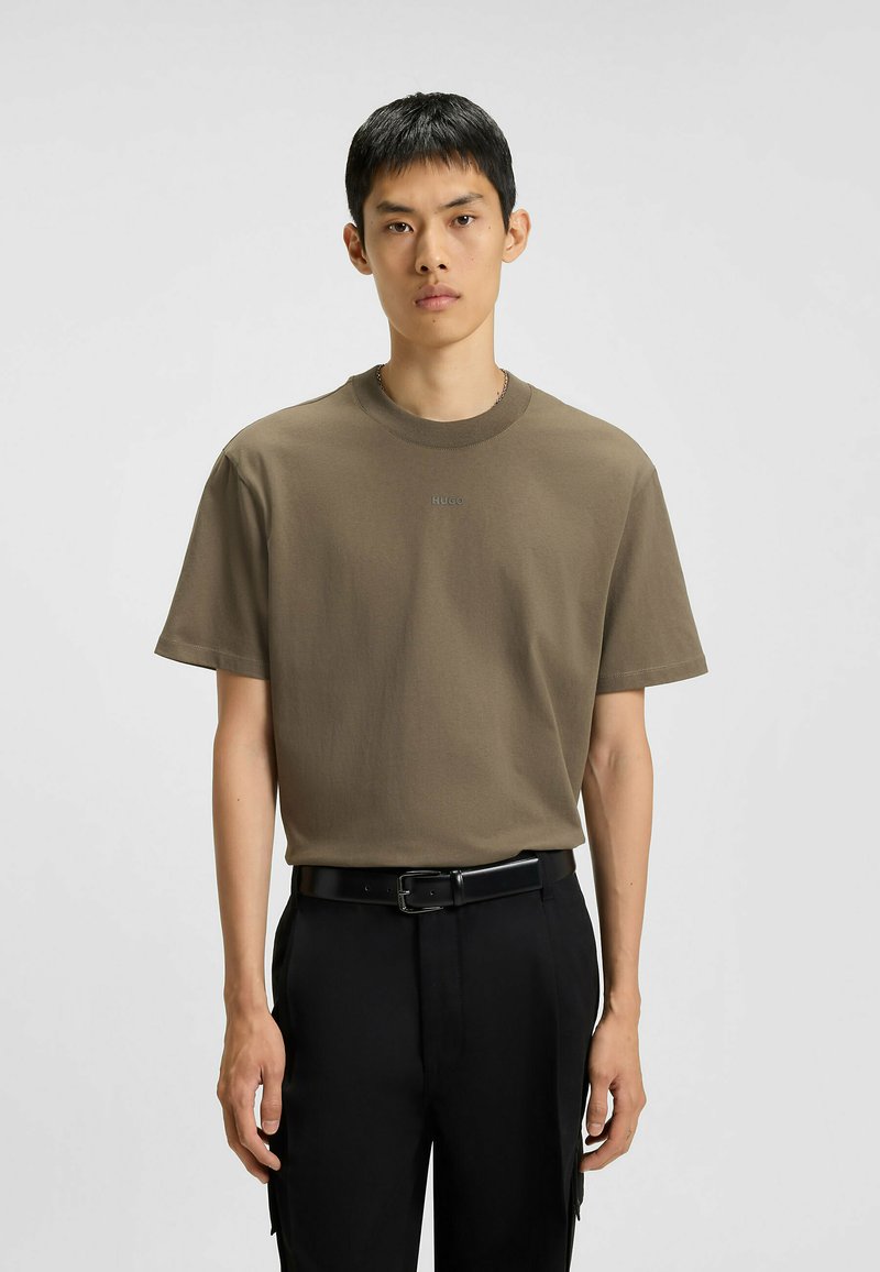 T-shirt in cotone marrone con scollo rotondo e maniche corte, caratterizzato da un sottile logo "HUGO" al centro. Indossato con pantaloni neri e una cintura.