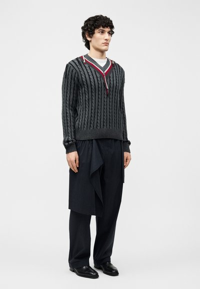 Jeune homme aux cheveux bouclés portant un pull en maille torsadée gris foncé avec fermeture éclair et un pantalon ample noir à rayures fines avec tissu superposé.