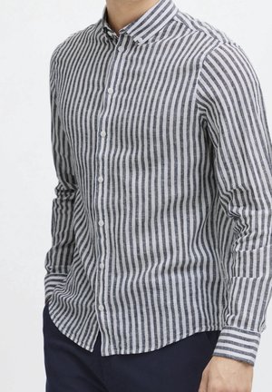Man wearing a long-sleeve, vertical black and white striped button-up shirt with dark pants, standing with one hand in pocket.