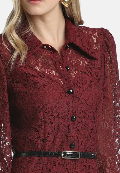 Burgunderfarbige Spitzenbluse mit Kragen, langen Ärmeln, schwarzen Knöpfen und einem schwarzen, taillierten Gürtel. Florales Spitzenmuster über die gesamte Bluse.
