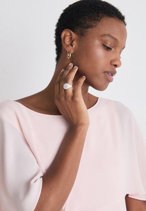 Silberring mit einem strukturierten, birnenförmigen Design und mehreren kleinen Edelsteinen, getragen an einer Hand mit einem goldenen Creolenohrring, vor einem sanften rosa Oberteil.