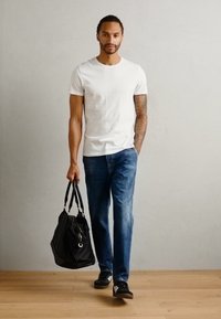 Geantă de voiaj neagră, tricou alb din bumbac, blugi din denim albastru, adidași negri cu accente albe. Podea din lemn, fundal neutru.