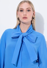 Blusa blu brillante con un grande fiocco al collo, realizzata in un tessuto morbido e liscio con maniche lunghe. Orecchini d'oro di grande impatto completano il look.