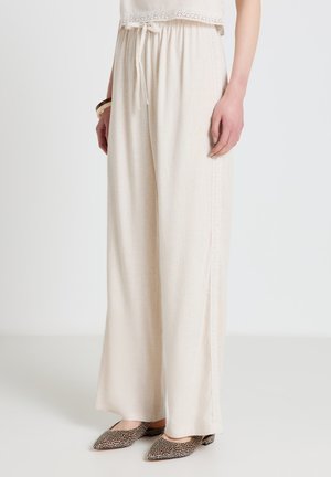 Frau trägt locker sitzende, cremefarbene weite Hosen mit Kordelzug in der Taille und beige spitze flache Schuhe mit Leopardenmuster.