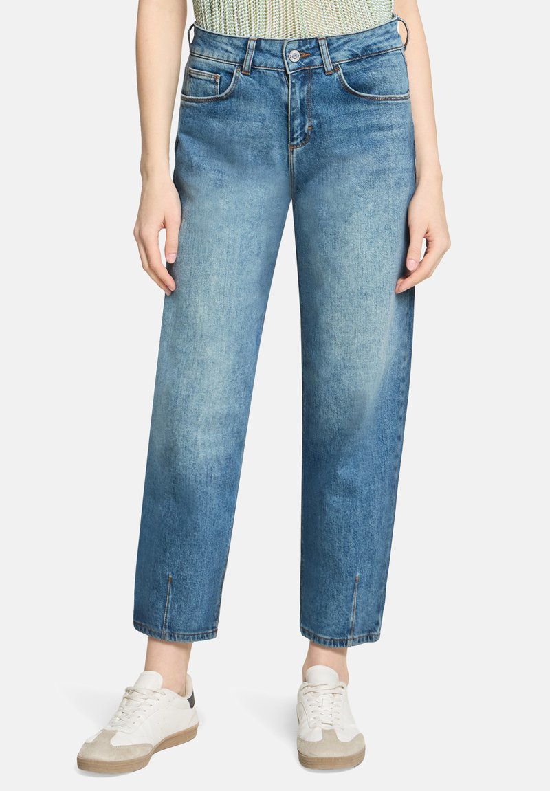 Kvinde iført lyseblå jeans med lige ben, forlommer og små slids foran på benene, kombineret med hvide sneakers.