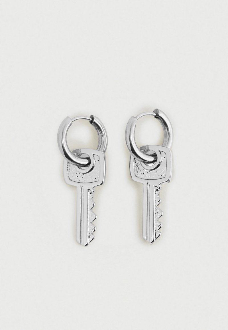 Pár stříbrných náušnic ve tvaru kruhu, z nichž každá má malý přívěsek ve tvaru klíče zavěšený na kruhu, na čistém bílém pozadí.