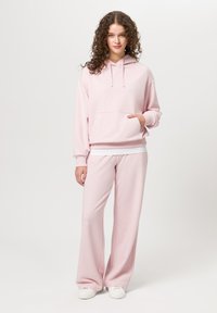 Růžová mikina s kapucí a přední kapsou, doplněná volnými kalhotami ve stejném stylu. Měkká tkanina s hladkým povrchem. Bílé tenisky.