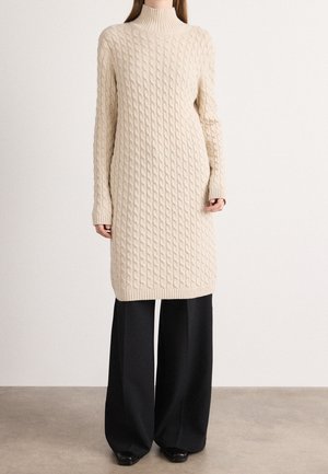 Personne portant une robe-pull à col roulé en maille torsadée beige sur un pantalon noir à larges jambes et des chaussures noires sur un fond uni.