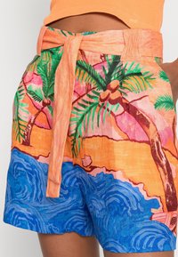 Shorts taille haute avec une ceinture en tissu orange, imprimé tropical présentant des palmiers, des vagues bleues et une palette de couleurs de coucher de soleil.