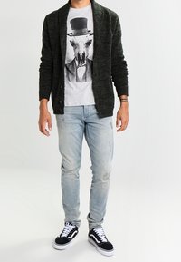 Mies, der einen schwarzen Cardigan, ein graues Grafik-T-Shirt mit einem Tierschädel und einem Zylinder, hellen verwaschenen Jeans und schwarzen Turnschuhen mit weißen Schnürsenkeln trägt.