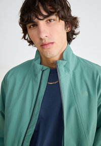 Lightweight, mint green jacket with a stand-up collar and zipper closure, made of smooth fabric. Worn over a navy shirt with a gold chain accent.