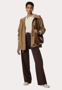 Giacca cappuccio color tan, maglione di cotone crema, pantaloni larghi marroni e borsa a spalla marrone. Gli accessori includono polsini a righe e orecchini.