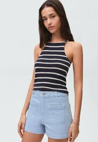 Prugasta pletena majica bez rukava v mornarsko modri in kremni barvi, v kombinaciji s svetlo modrimi jeans kratkimi hlačami, ki imajo dva sprednja žepa in gumbasto zaponko.
