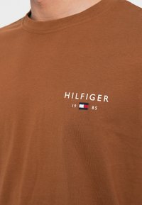 Καφέ βαμβακερό μπλουζάκι με στρογγυλή λαιμόκοψη, που διαθέτει κείμενο "HILFIGER" και μια μικρή σημαία σε κόκκινο, λευκό και μπλε χρώμα στο στήθος.